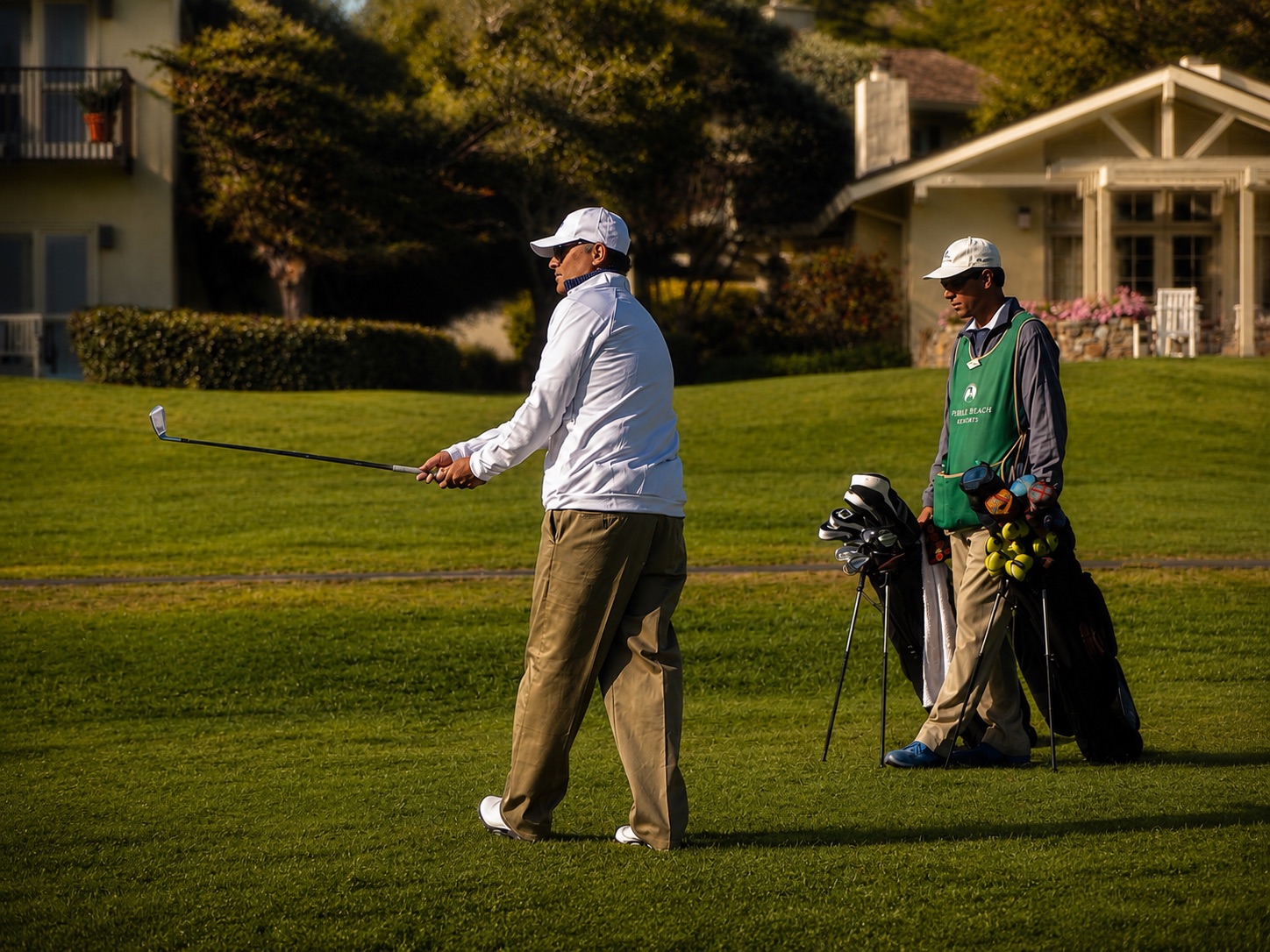 Hunt finishing a golf swing on the fairway at Pebble Beach in warm evening light, his caddie waiting with the bag beside him.